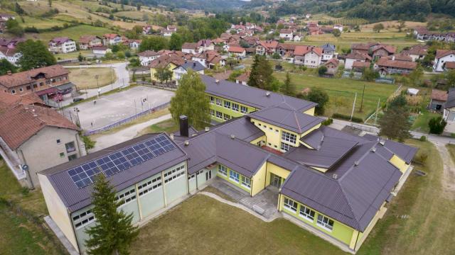 Aerial view of a school with solar panels on its roof, surrounded by houses and green hills