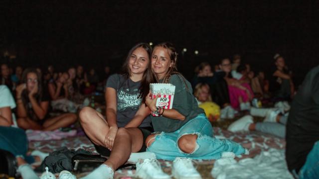 Two young women smiling on a blanket at an open-air cinema at night, one holding popcorn