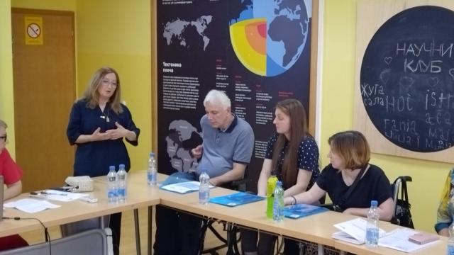Six people at a meeting table in a room with a science poster as a woman stands and speaks