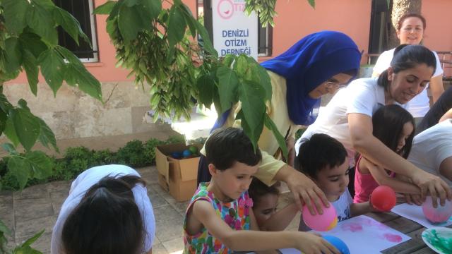 Children paint with balloons at an outdoor table, helped by women from the Denizli Autism Association