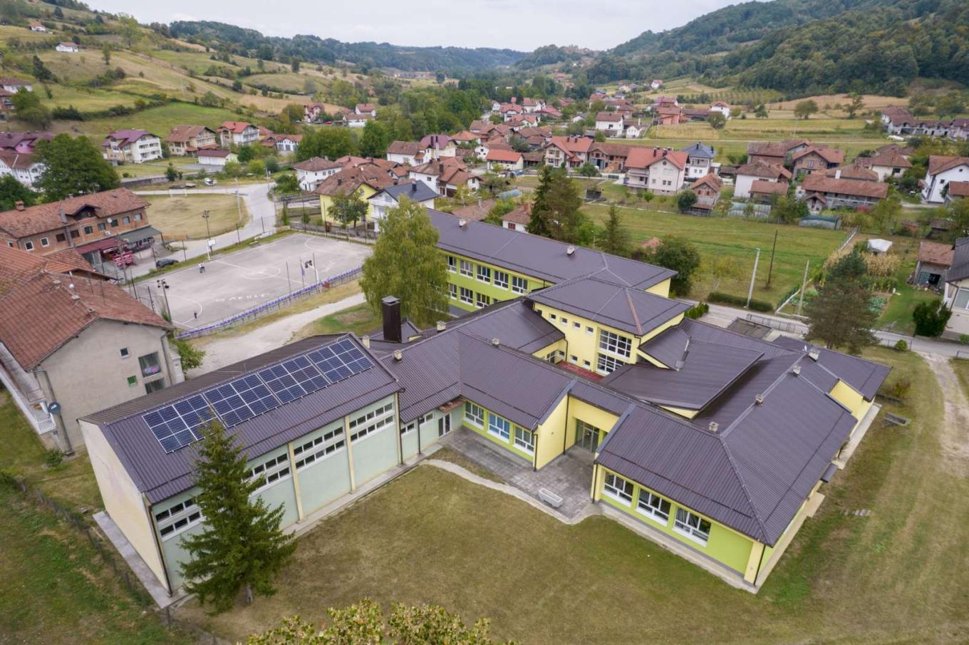 Aerial view of a school with solar panels on its roof, surrounded by houses and green hills