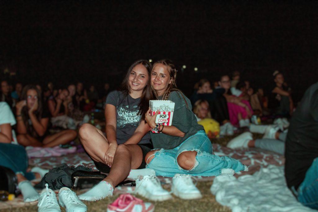 Two young women smiling on a blanket at an open-air cinema at night, one holding popcorn