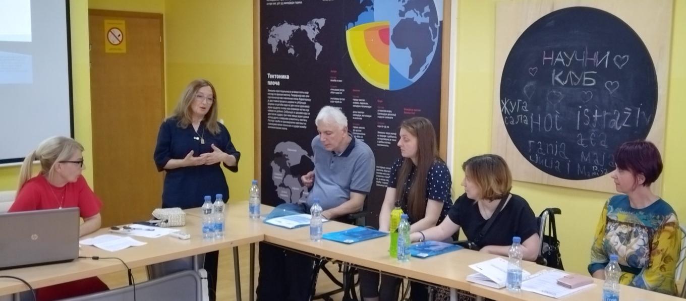 Six people at a meeting table in a room with a science poster as a woman stands and speaks