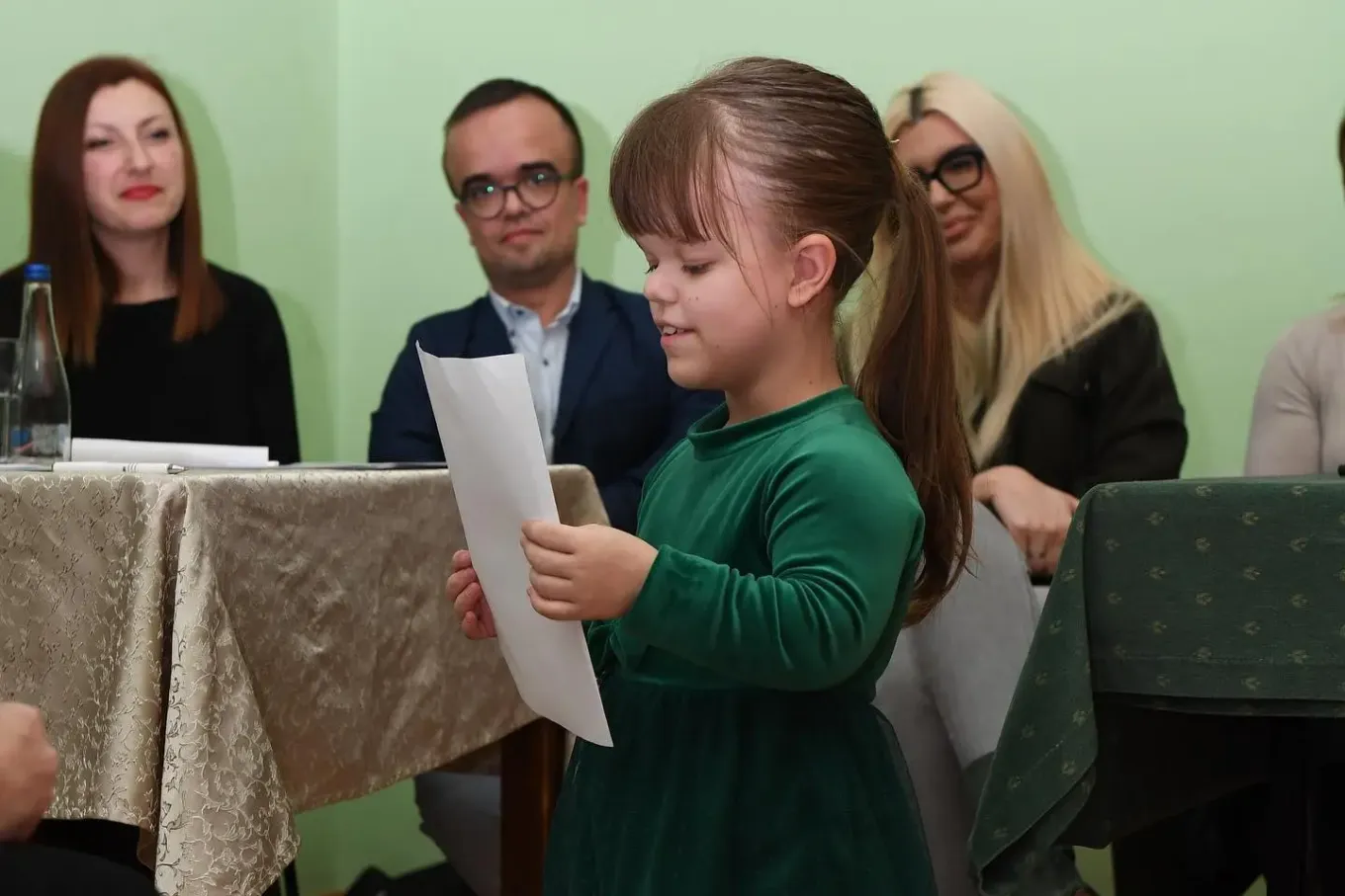 A young girl reads aloud from a sheet of paper at an event, with adults seated behind her