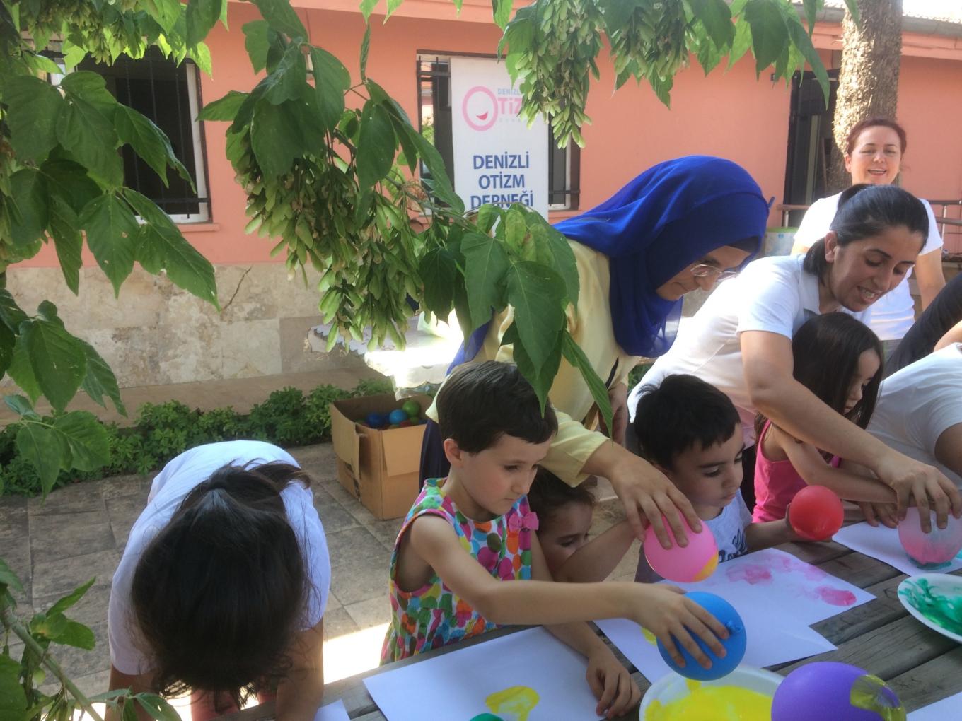 Children paint with balloons at an outdoor table, helped by women from the Denizli Autism Association